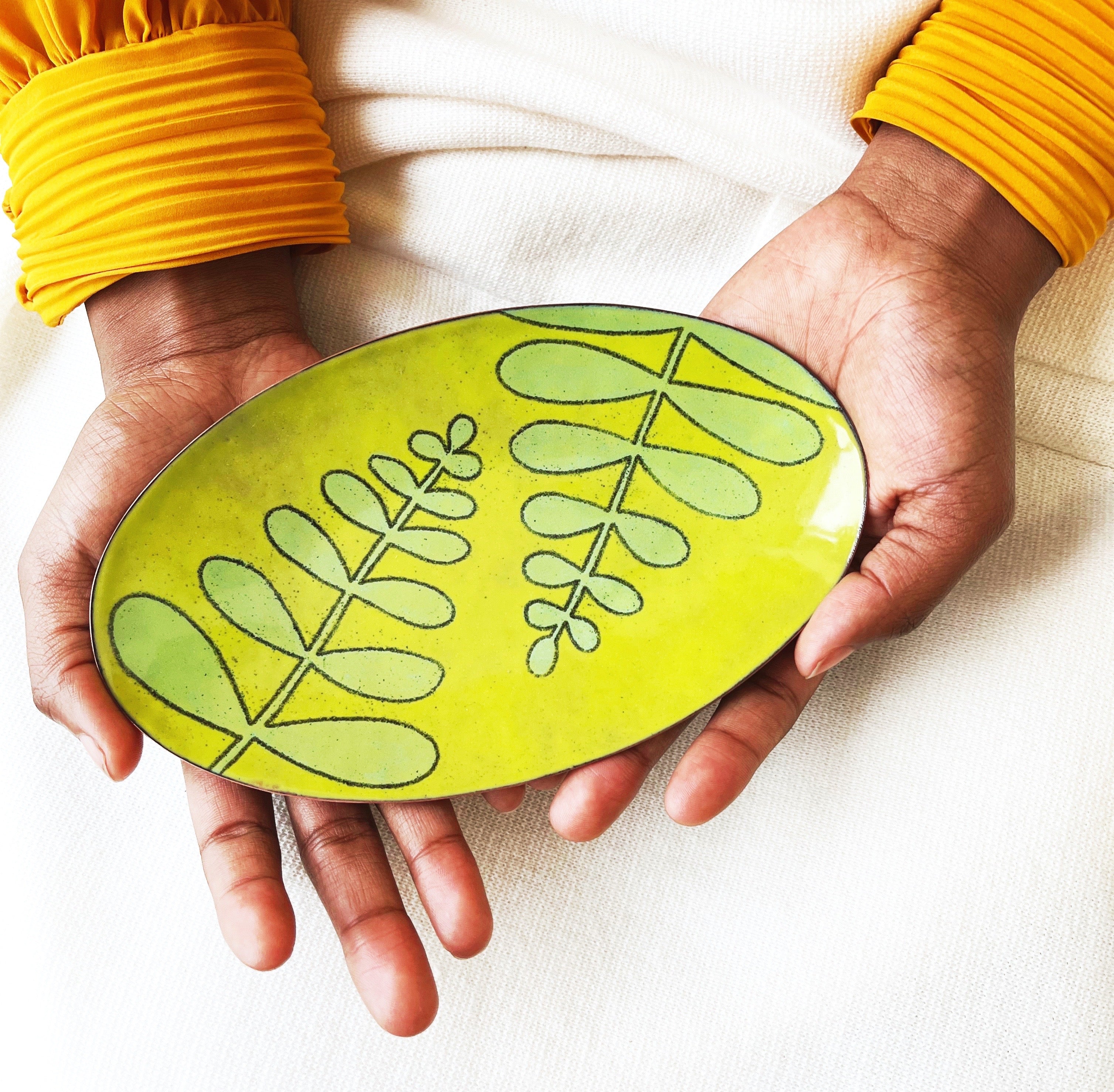 A black woman in a gold shirt holds a handmade dish. It is chartreuse with a mint green fern pattern drawn on. It is layered glass enamel, kiln fired on copper.