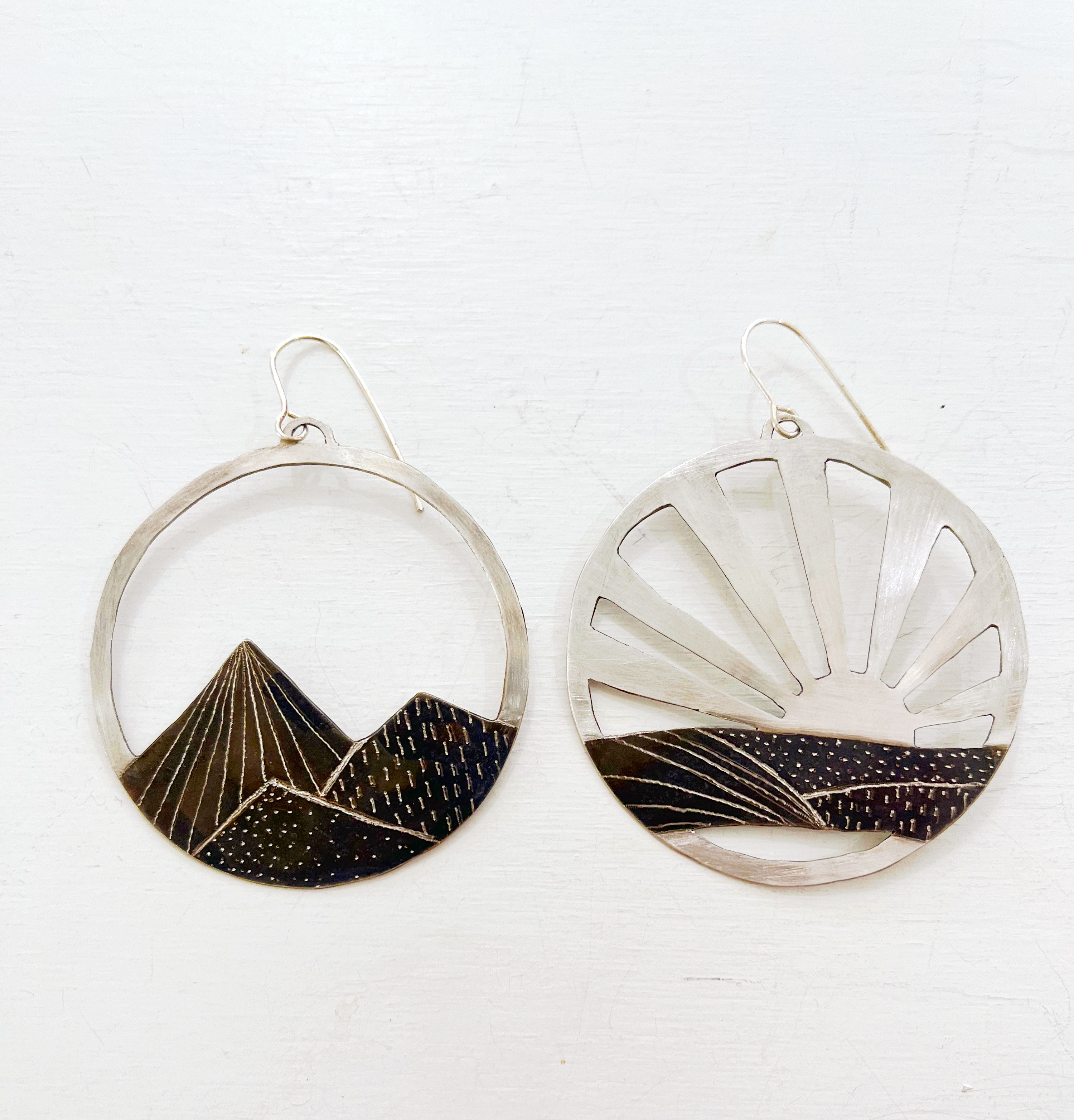 Hand cut sterling silver hoop Earrings. One is of a mountain and one of a sunrise. They are a set. the landscape has a patina and is hand drawn. 